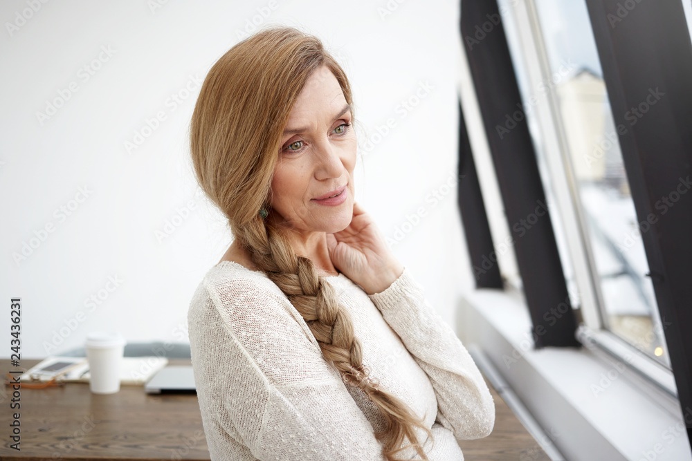 Attractive green eyed European middle aged female in her sixties standing at panoramic window, touching her neck, having pensive thoughtful facial expression, deep in thoughts, enjoying leisure time Attractive green eyed European middle aged female in her sixties standing at panoramic window, touching her neck, having pensive thoughtful facial expression, deep in thoughts, enjoying leisure time