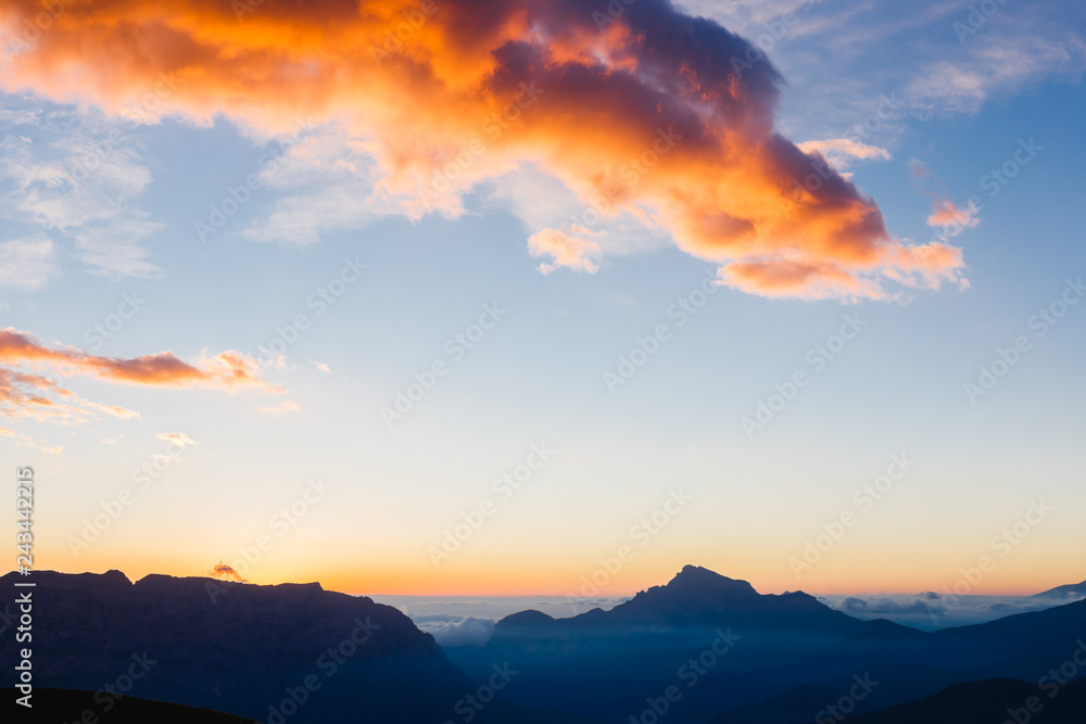 Colorful orange clouds in the big blue sky over the dark mountains at sunrise. Beautiful morning landscape in the Caucasus Mountains. Republic of North Ossetia–Alania, Russia