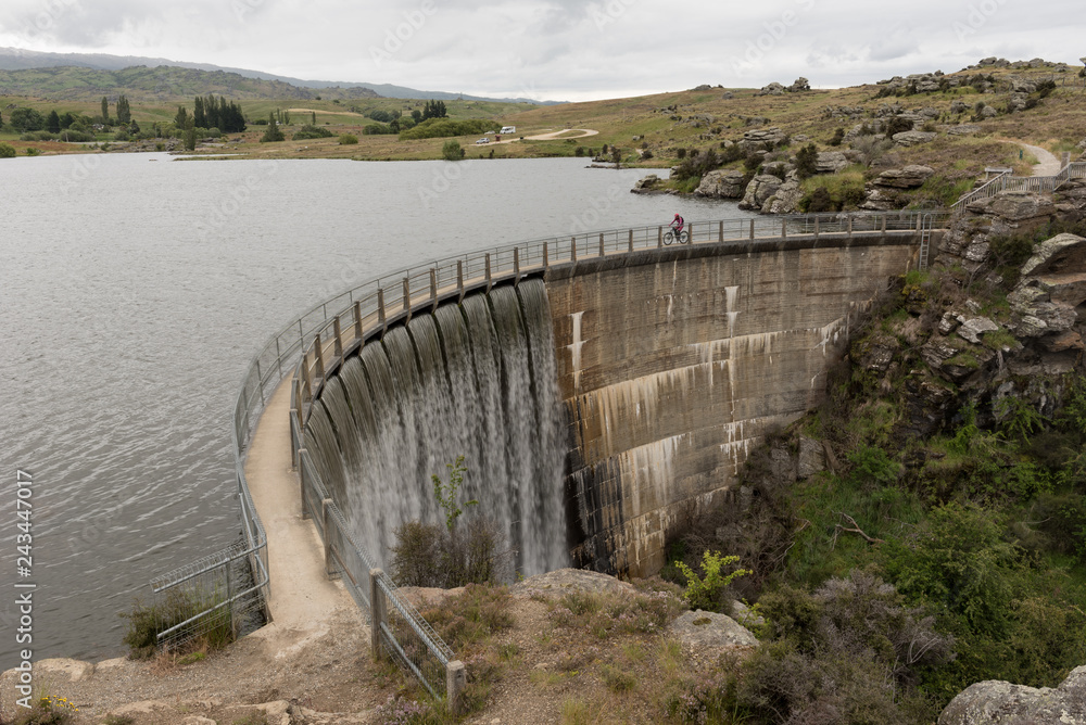 A cyclist crossing Butcher's Dam, Flat Top Hill Conservation Area, releasing excess water over its spillway. Central Otago, New Zealand.