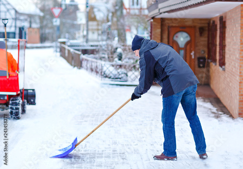 Man with snow shovel cleans sidewalks in winter during snowfall. Winter time in Europe. Young man in warm winter clothes. Snow and weather chaos in Germany. Snowstorm and heavy snowing. Schneechaos