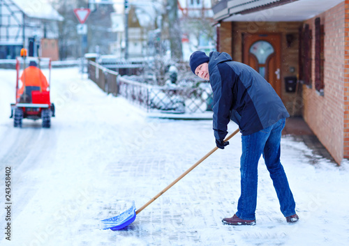 Man with snow shovel cleans sidewalks in winter during snowfall. Winter time in Europe. Young man in warm winter clothes. Snow and weather chaos in Germany. Snowstorm and heavy snowing. Schneechaos