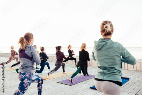 Young girl yogi instructor doing morning yoga class with group of people outdoor on wooden terrace by the sea. Rear view, selective focus on the woman teacher. Horse rider exercise, anjaneyasana pose photo