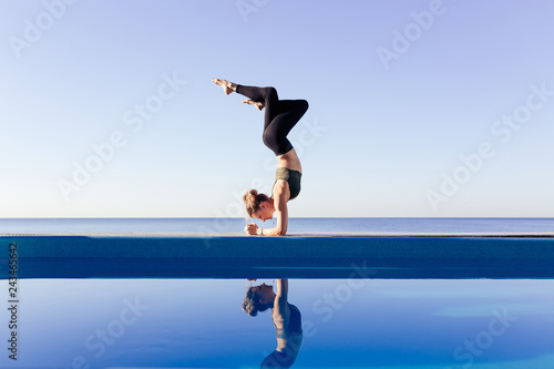 Young attractive woman practicing yoga. Headstand, Garuda salamba sirsasana, working out by pool, above the beach, relaxing against blue sky. Healthy, beauty concept, reflection on water, meditating photo