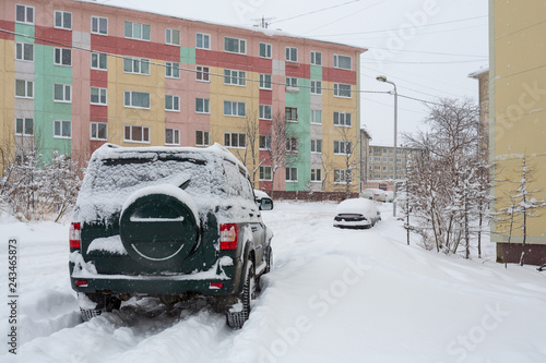Off-road car on a snowy road in the yard after a snowfall. Around the snowdrifts and a lot of snow on the machines. Multi-colored panel buildings. Cold winter weather. Magadan, Far East of Russia.