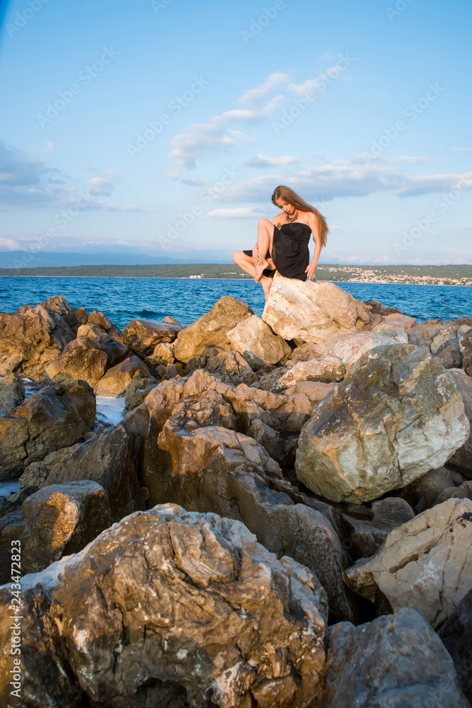 A beautiful woman in an elegant black dress sitting and posing on the rocks next to the sea on a sunny day.