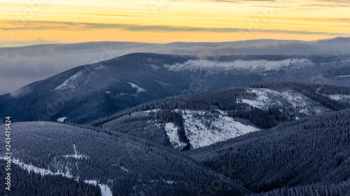 Beautiful view of wooden and snowy mountains partially covered in clouds with colorful sky, viewed from Horni Mala Upa in Krkonose (Giant Mountains) in the direction of Pec pod Snezkou