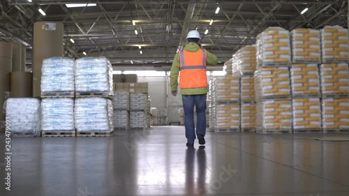 Worker in hard hat and orange uniform walking and talking by cellphone in warehouse - back view photo