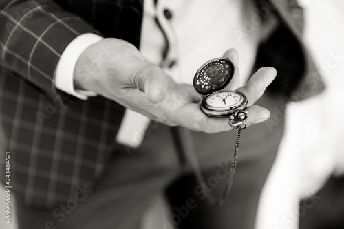 A man in a checkered suit holds an open pocket watch in his hand, the dial is visible. Black and white close up photo