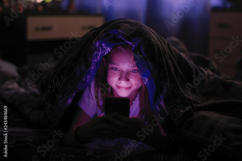 Little girl at night looks at smartphone under a blanket in bed.