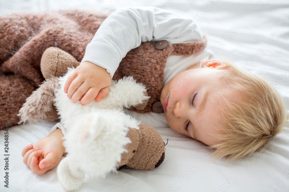 Sweet baby boy in cute overall, sleeping in bed with teddy bear stuffed toys, winter time