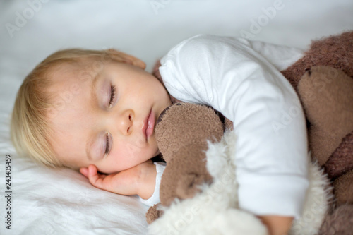 Sweet baby boy in cute overall, sleeping in bed with teddy bear stuffed toys, winter time