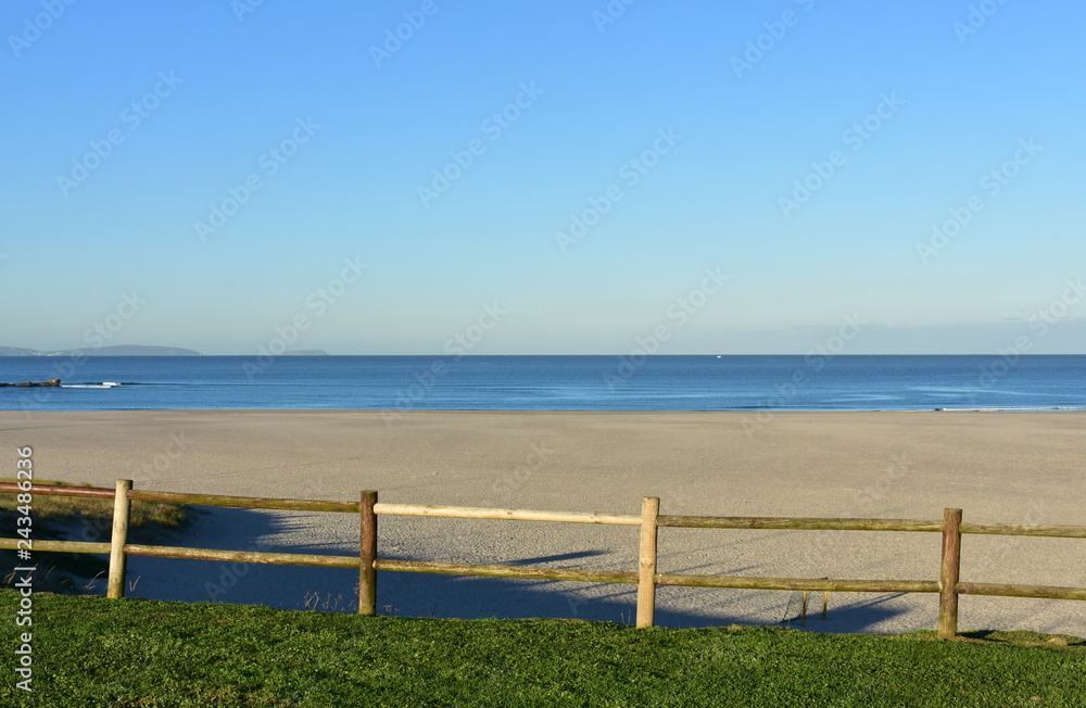 Fototapeta premium Beach with grass and wooden fence. Morning light, sunny day, blue sky. Galicia, Coruna, Spain.