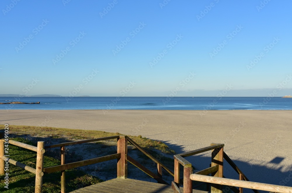 Beach with grass and wooden boardwalk with fence . Morning light, sunny day, blue sky. Galicia, Coruna, Spain.