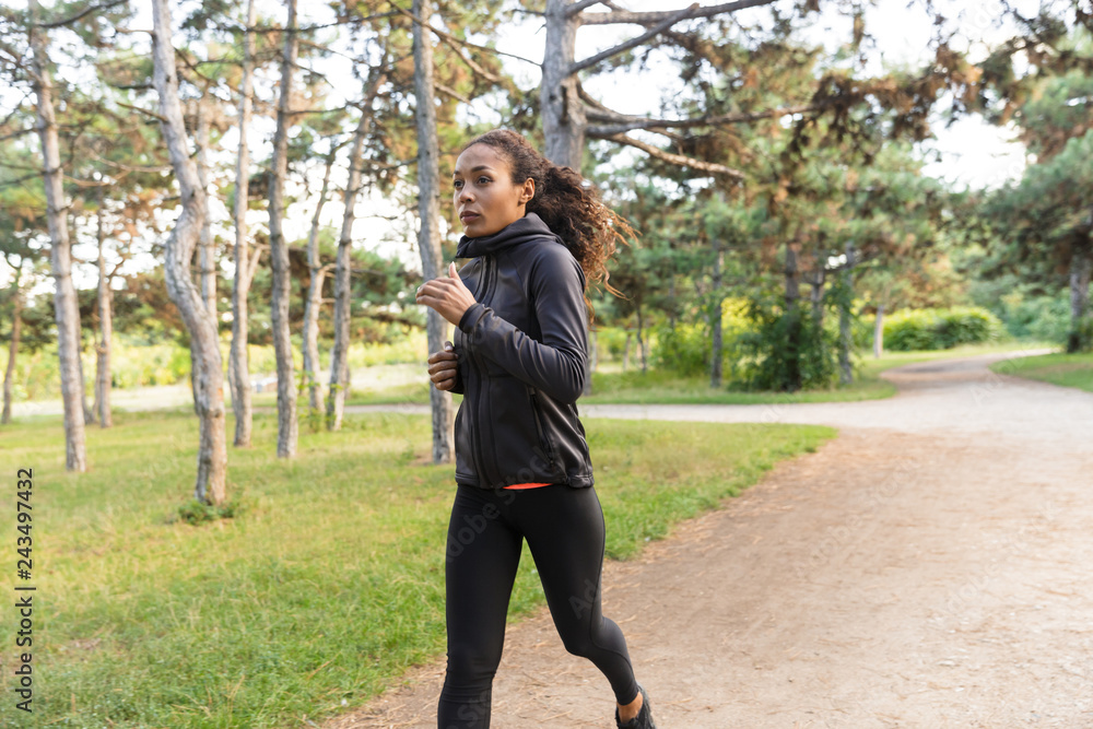 Image of brunette woman 20s wearing black tracksuit working out, while running through green park
