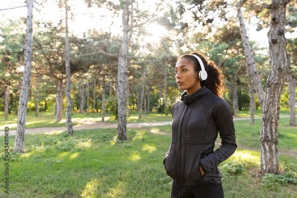 Image of young woman 20s wearing black tracksuit and headphones, walking through green park Image of young woman 20s wearing black tracksuit and headphones, walking through green park