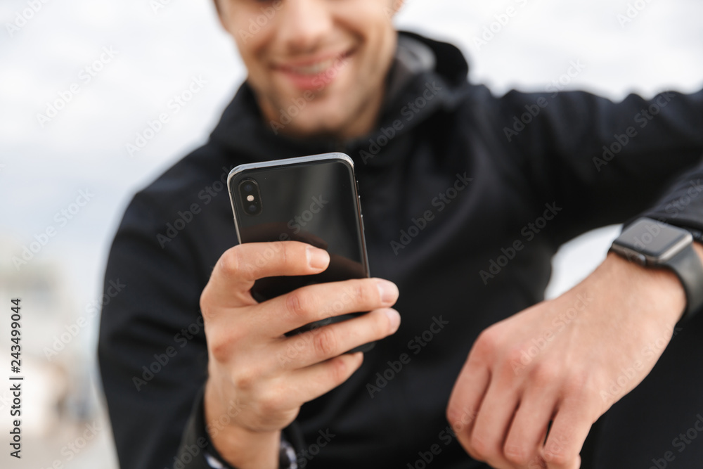 Image of mature sportsman 30s in black sportswear, using smartphone while sitting on boardwalk at seaside Image of mature sportsman 30s in black sportswear, using smartphone while sitting on boardwalk at seaside
