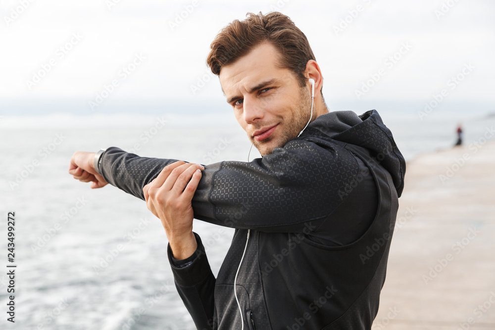 Fototapeta premium Image of european sportsman 30s in black sportswear and earphones, working out at seaside