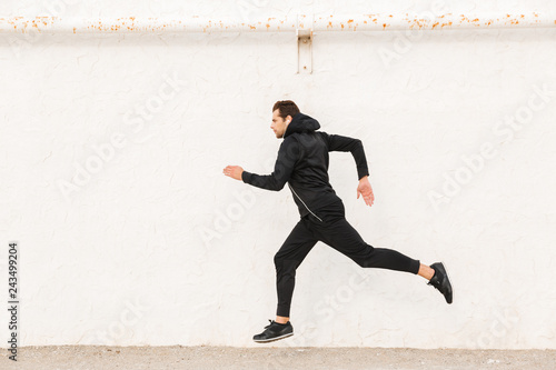 Image of caucasian man 30s in black sportswear and earphones, running along white wall outdoor