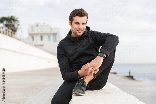 Image of sportive adult man 30s in black sportswear, sitting on boardwalk at seaside