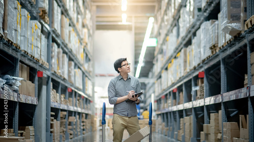 Young Asian man worker doing stocktaking of product in cardboard box on shelves in warehouse by using digital tablet and pen. Physical inventory count concept