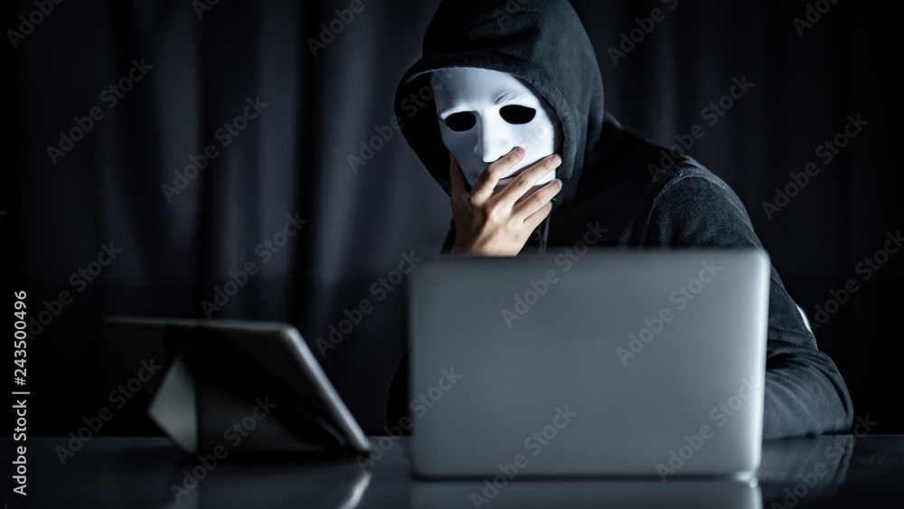 Mystery male hoodie hacker holding white mask looking at laptop computer. Anonymous social masking. Ransomware cyber attack or internet security concepts Mystery male hoodie hacker holding white mask looking at laptop computer. Anonymous social masking. Ransomware cyber attack or internet security concepts
