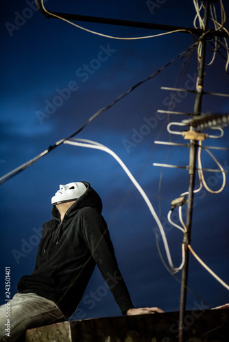 Mystery hoodie man in white mask sitting with antenna on rooftop of abandoned building looking up at the sky during the twilight time. Bipolar disorder or Major depressive disorder. Depression concept