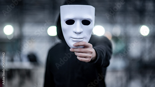 Mystery hoodie man with black mask holding white mask in his hand. Anonymous social masking or bipolar disorder concept.