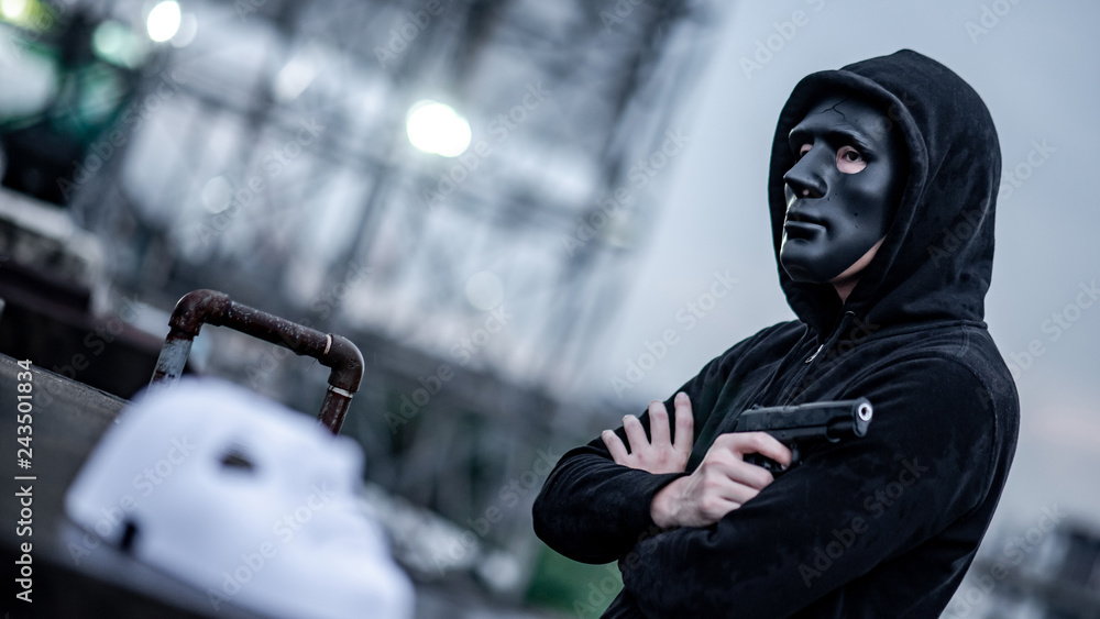 Mystery hoodie man in broken black mask holding gun feeling guilty. Crime and violence concepts Mystery hoodie man in broken black mask holding gun feeling guilty. Crime and violence concepts