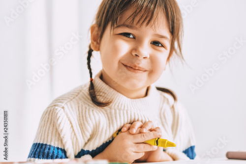Closeup portrait of beautiful happy little girl paints with oil pencils, sitting at white desk at home. Pretty smiling preschool kid posing with yellow pencil. People, childhood, education concept
