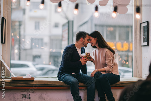 Loving couple dressed in sweaters and jeans is sitting close to each other on the windowsill in a cafe and holding cups in their hands