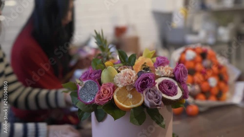 A close-up scene of a bouquet of flowers which consist of violet roses, green leaves, half of orange, dragon fruit. This quaint bouquet in a round-shaped box is made by a two blurred florists. photo