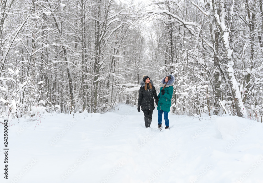 Two caucasian girls are walking in the winter forest during a snowfall. The happy young womens are holding hands in rural.