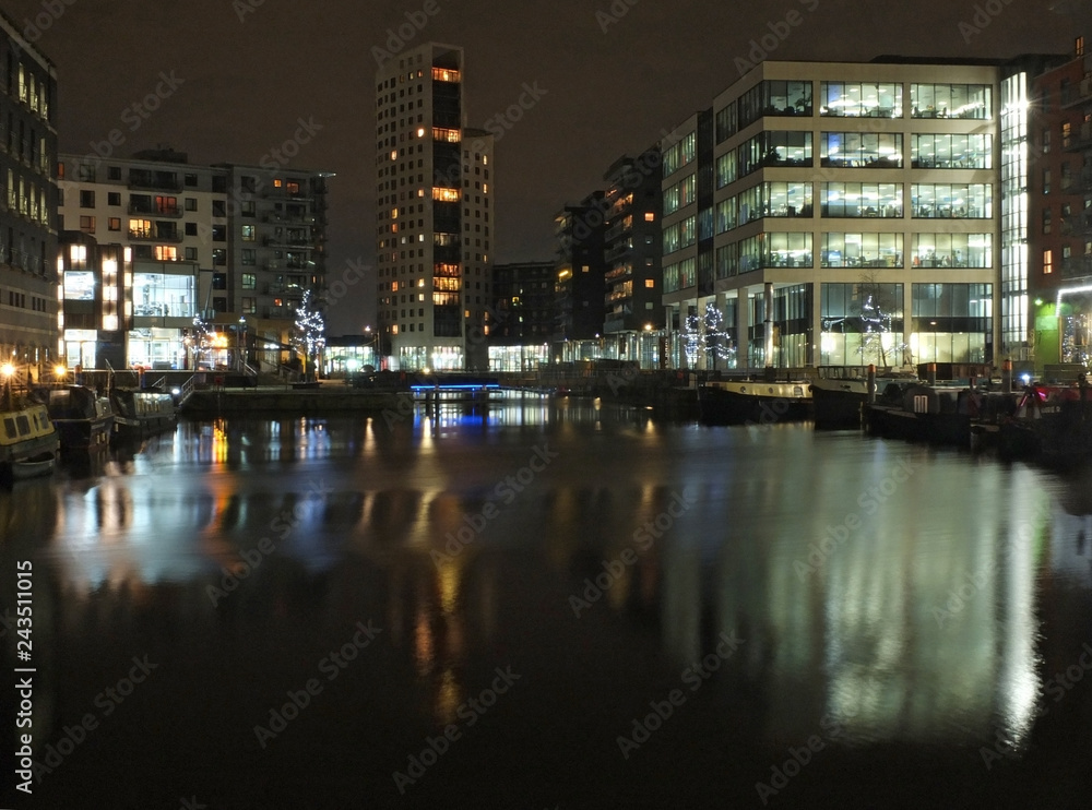 clarence dock in leeds at night with brightly illuminated buildings reflected in the water and boats moored along the sides