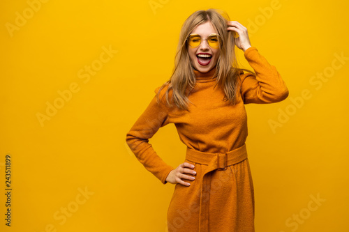 Excited young woman in orange dress, widely smiling, looking at camera. Isolated on yellow background