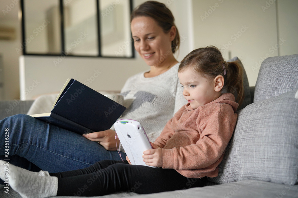 Fototapeta premium Mother and young daughter reading and playing on tablet at home