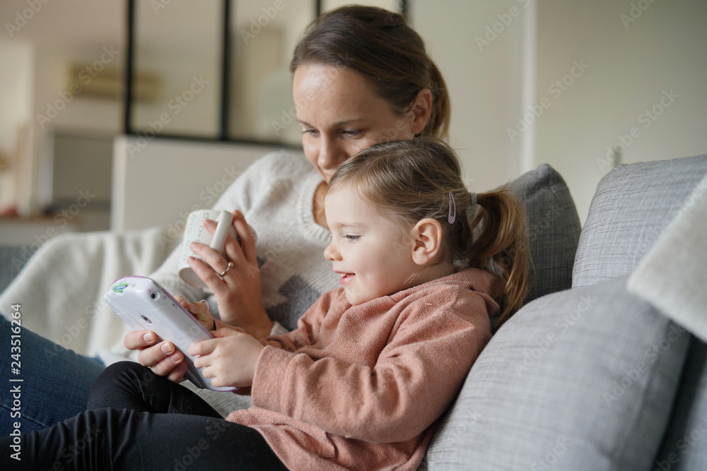 Mother and young daughter playing games on child's tablet at home - obrazy, fototapety, plakaty 