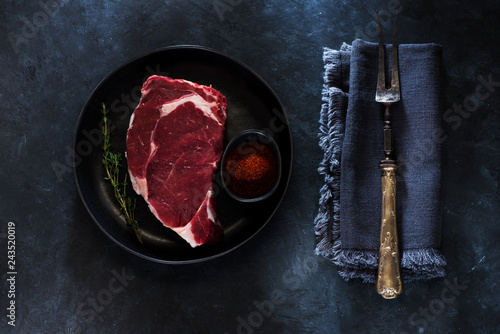 Raw Steak Ribeye in plate, meat fork, herbs and spices on a dark concrete background