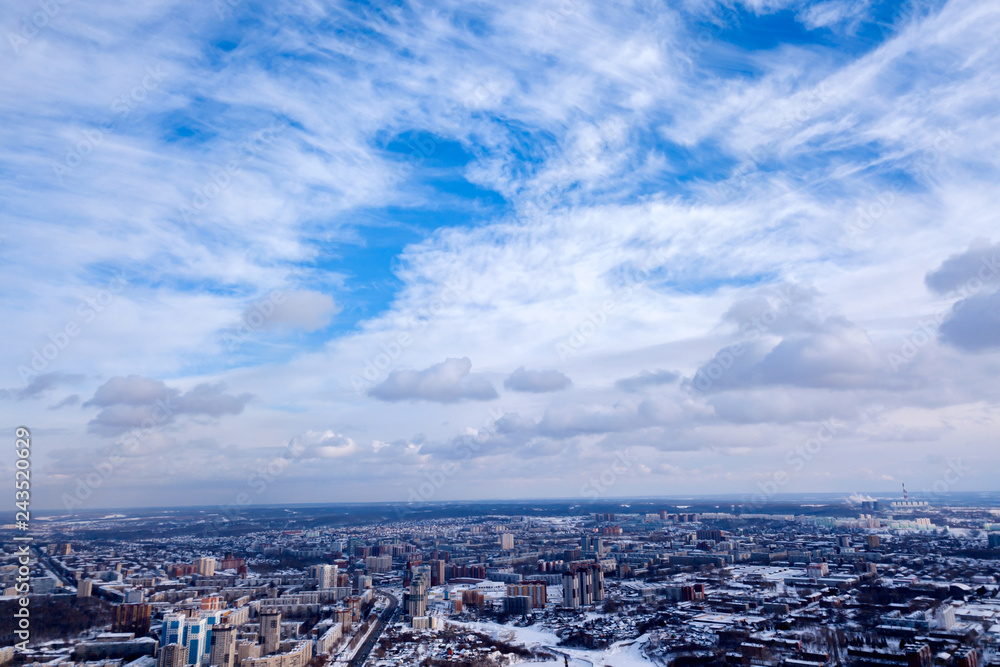 Scenic picture of the city in winter of a aerial, clear blue sky with white clouds, high-rise houses with roofs under the snow, streets with trees, industrial plants in the distance.