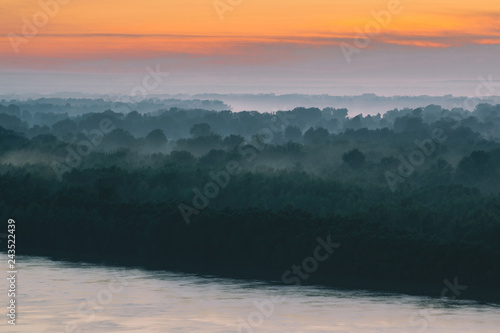 Mystical view on riverbank of large island with forest under haze at early morning. Mist among layers from tree silhouettes under warm predawn sky. Morning atmospheric landscape of majestic nature. © Daniil