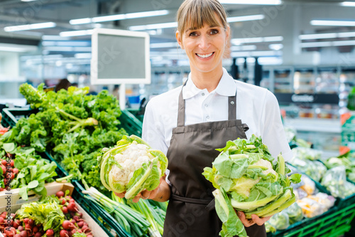 Quadro su tela Shop assistant woman in supermarket showing the fresh produce with pride looking