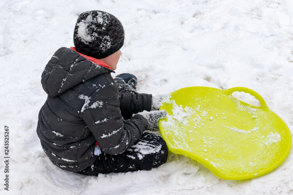 Φωτογραφία A boy of seven years old sitting on the snow and a green plastic saucer sled lying near him