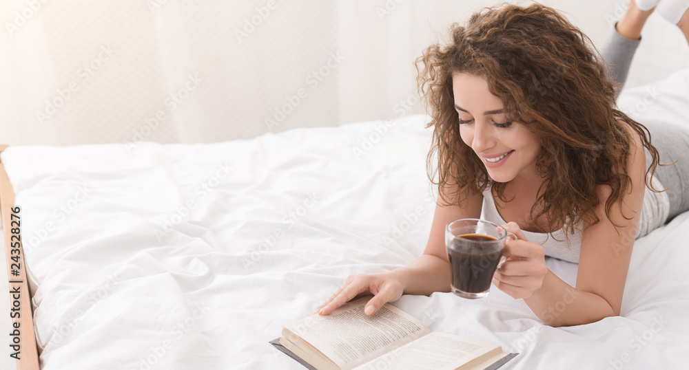 Cheerful girl reading book and drinking coffee in bed
