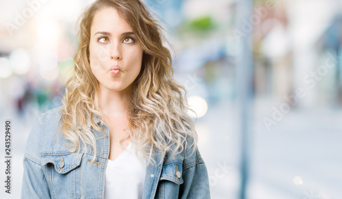 Beautiful young blonde woman wearing denim jacket over isolated background making fish face with lips, crazy and comical gesture. Funny expression.
