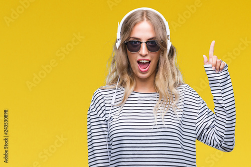 Beautiful young blonde woman wearing headphones and sunglasses over isolated background pointing finger up with successful idea. Exited and happy. Number one.