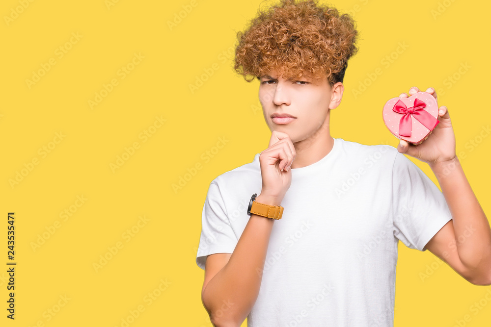 Young handsome man holding heart box as gift of valentine's day serious face thinking about question, very confused idea Young handsome man holding heart box as gift of valentine's day serious face thinking about question, very confused idea