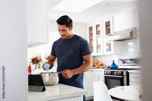 Millennial Hispanic man preparing cake mixture in kitchen, following a recipe on a tablet computer