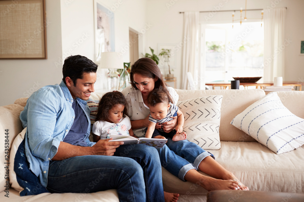 Hispanic couple sitting on the sofa reading a book at home with their baby son and young daughter Hispanic couple sitting on the sofa reading a book at home with their baby son and young daughter