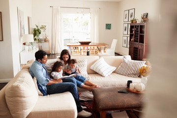 Young Hispanic family sitting on sofa reading a book together in the living room, seen from doorway