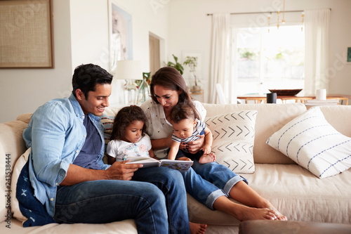 Hispanic couple sitting on the sofa reading a book at home with their baby son and young daughter photo