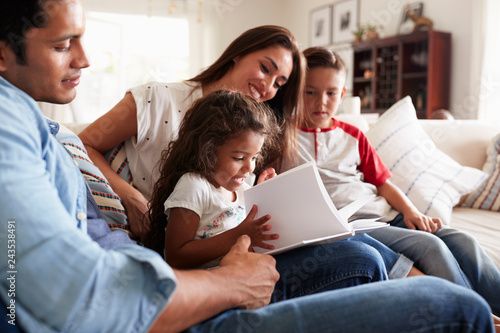 Fotografía Young Hispanic family of four sitting on the sofa reading book together in their